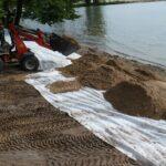 Prace budowalne przy budowie Eko Plaży Giżycko. Zdjęcie przedstawia nasypywanie piasku pod przyszłą plażę. Na zdjęciu widać brzeg jeziora Niegocin, piasek wysypywany na podkład oraz czerwoną małą wywrotkę.